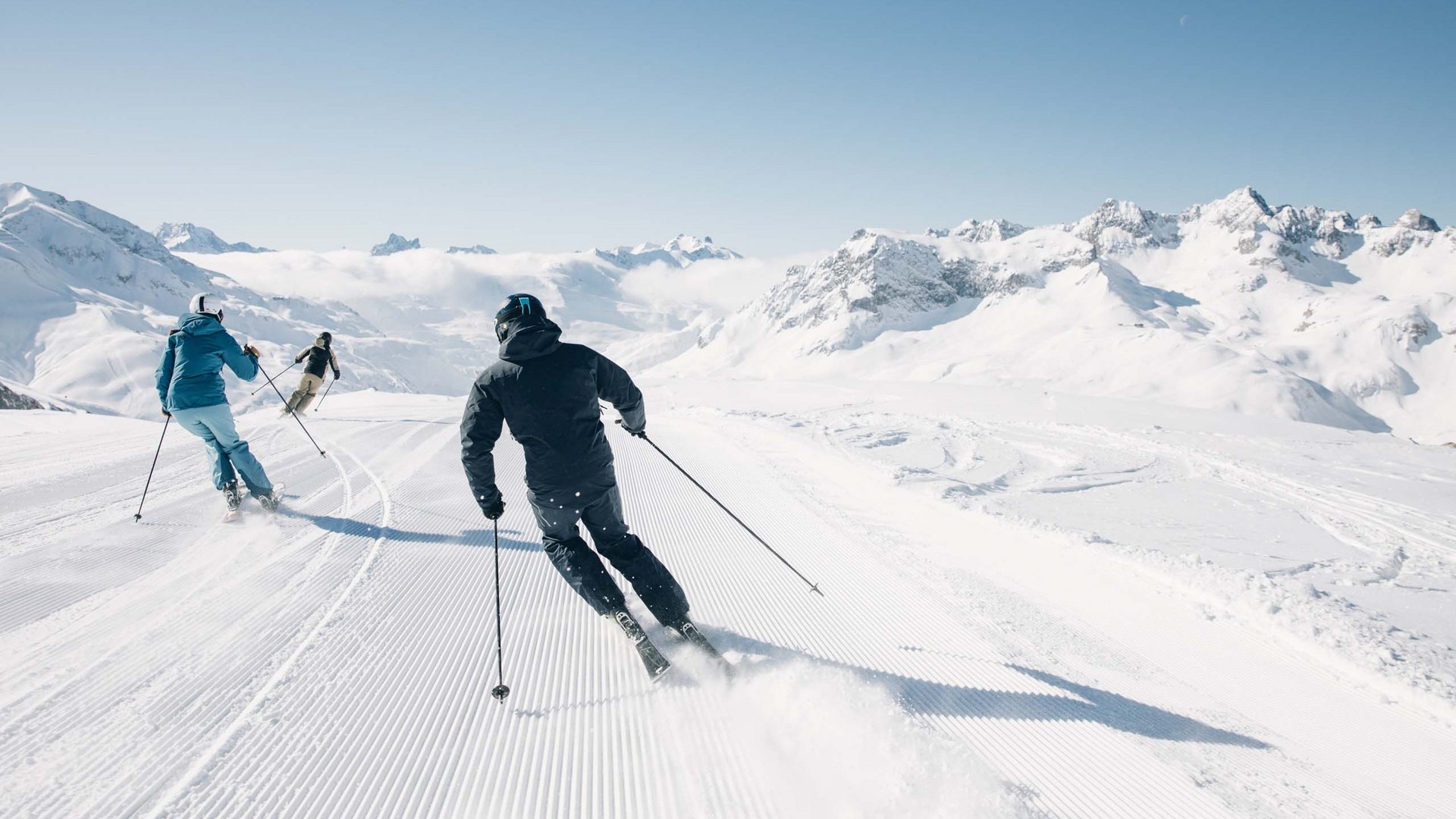 Three skiers skiing down a groomed slope in snowy mountains