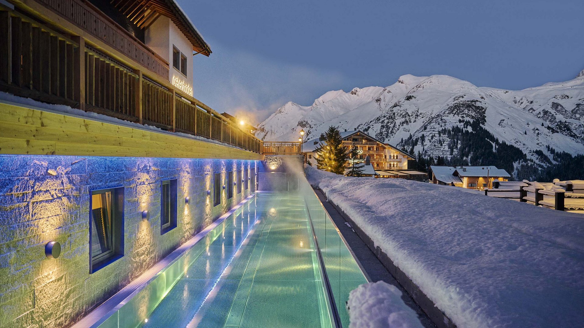 A ski-in, ski-out hotel in Oberlech with pool and spa Lit swimming pool beside snowy building with Alps mountains in the background at dusk